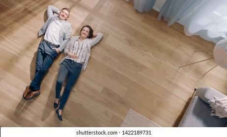 Young Couple Are Lying On A Wooden Flooring In An Apartment. They Are Happy, Smile And Laugh. Cozy Living Room With Modern Interior, Grey Sofa And Wooden Parquet. Top View Camera Shot.