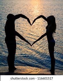 young couple making heart shape with arms on beach against golden sunset