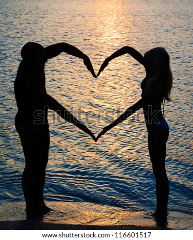 young couple making heart shape with arms on beach against golden sunset
