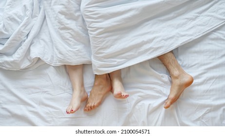 Young Couple Making Love After Waking Up Lying On Soft Bed Sticking Bare Legs Out Of White Sheet In Early Morning At Home, Upper View.