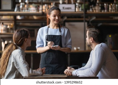 Young couple making order in cafe, attractive smiling waitress serving customers, writing in notepad, coffeehouse female worker talking with man and woman about menu, offering food and drinks - Powered by Shutterstock