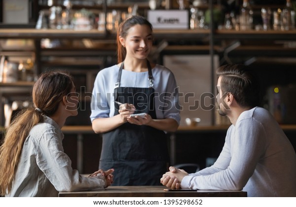 カフェで注文を取る若い夫婦 笑顔の魅力的なウエートレス メモ帳で書く 女性と男女でメニューを話す 飲食物を提供する 喫茶店の女性労働者 の写真素材 今すぐ編集
