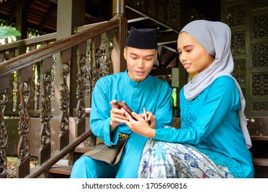 Young couple of malay muslim in traditional costume watching online content in a smart phone during Eid al-Fitr celebration at wooden stair.