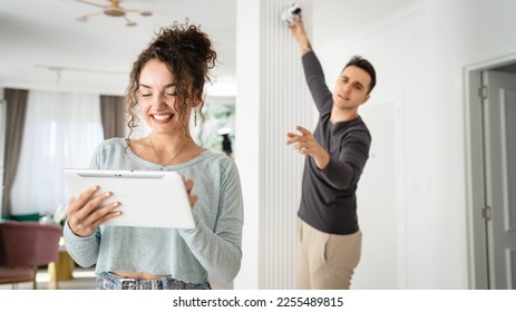 Young couple man and woman husband and wife or caucasian boyfriend and girlfriend setting up home surveillance security camera cctv protecting their home apartment bright room real people copy space - Shutterstock ID 2255489815