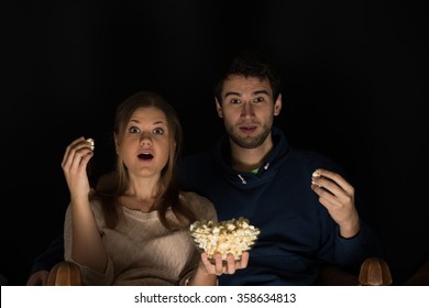 Young Couple, Man And Woman, Sitting In The Dark Room In The Front Of Tv Watching Movie And Eating Popcorn, Showing Emotions