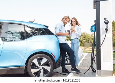 Young couple man and woman traveling together by new car having stop at charging station boyfriend holding cup drinking hot coffee talking with girlfriend browsing smartphone smiling joyful while