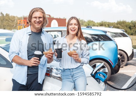 Young couple man and woman traveling by electric car having stop at charging station standing holding cups drinking hot coffee eating hamburgers looking camera laughing cheerful while having car fully