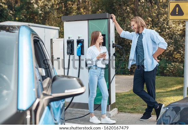 Young couple man and woman traveling by electric\
car having stop at charging station talking browsing smartphone\
looking at each other smiling happy while waiting for vehicle get\
fully charged