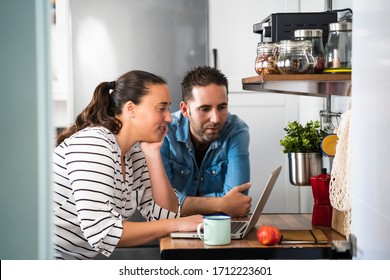 Young couple of man and woman using their laptop in the kitchen of their apartment. Work at home and stay home safely