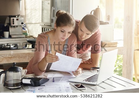 Young couple managing finances, reviewing their bank accounts using laptop computer and calculator at modern kitchen. Woman and man doing paperwork together, paying taxes online on notebook pc