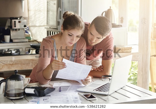 Young couple managing finances, reviewing their bank\
accounts using laptop computer and calculator at modern kitchen.\
Woman and man doing paperwork together, paying taxes online on\
notebook pc