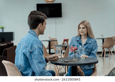 Young couple in matching denim jackets sitting at a modern lounge table, having a casual conversation over water glasses in a stylish indoor setting