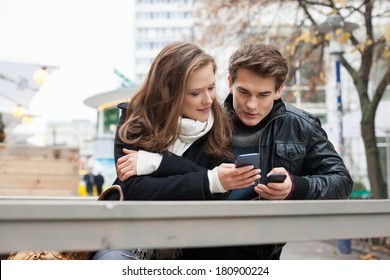 Young Couple Messaging Through Cell Phone While Leaning On Bench