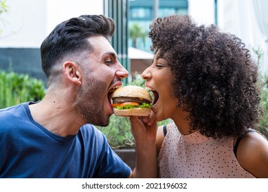 Junge, mehrethnische Liebhaber beißen den gleichen Käseeburger, zwei Freunde, die sich amüsieren, das gleiche Brot essen, ungesunde und Junk-Essen im Restaurant, Liebeskonzept