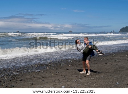Young couple on the beach by the waters edge on a sunny day with active waves and the man has picked the girl up in his arms as if to throw her in the sea and they are both laughing
