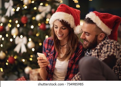 Young Couple On Christmas Eve Looking At Cell Phone Together 