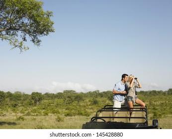 Young Couple On Safari Standing In Jeep And Looking Through Binoculars