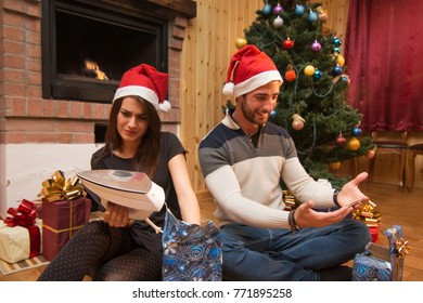 Young Couple Opening Christmas Gifts Under The Xmas Tree Wearing Santa Hats. Woman Unhappy With Bad Christmas Gift