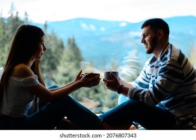 A young couple over a cup of coffee discussing everyday affairs, a guy and a girl sitting on the windowsill on a background of mountains and drinking coffee, relaxing in the Carpathians.
