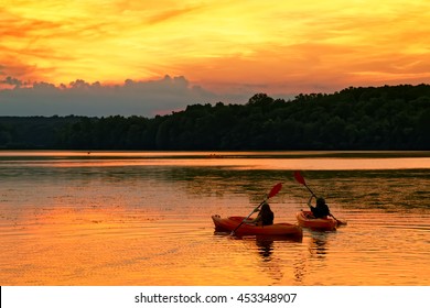 A Young Couple Paddle Kayaks On A Lake At Sunset.