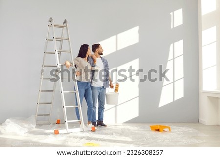 Young couple painting walls in their new home. Happy husband and wife with paint rollers renovating and decorating room in their house. Couple standing, hugging and looking at window in new apartment Foto stock © 