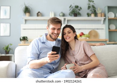 Young couple in pajamas watching online content in a smart phone sitting on a sofa at home in the living room