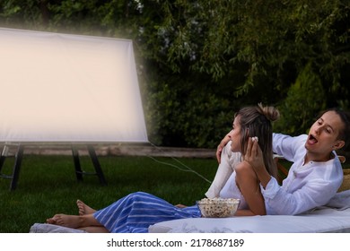 Young Couple In The Park Watching A Summer Movie. Boyfriend Stealing Opcorns From Behind To His Girlfriend