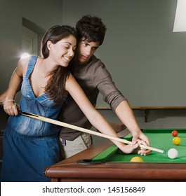 Young Couple Playing Pool In A Bar While Having A Night Out In Town With Young Man Teaching Woman How To Play, Smiling.