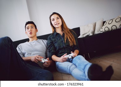 Young Couple Playing Video Games Together While Sitting In Their Living Room. Mixed Race Teenage Couple Holding Video Game Console Sitting On Floor Looking Away.