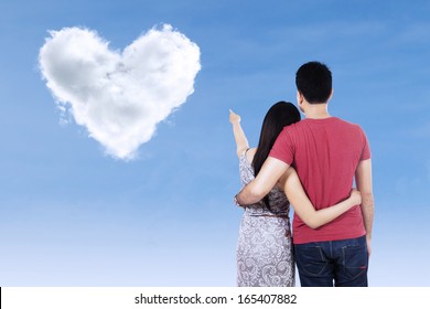 Young Couple Pointing At Clouds Shaped Of Heart On Blue Sky