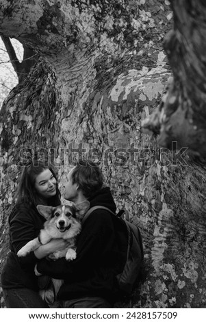 A young couple posing with cute dog welsh corgi pembroke, exchanging loving glances. The dog is perched in the mans arms, looking sweetly. Backdrop features a trunk of an old tree. Trendy monochrome.