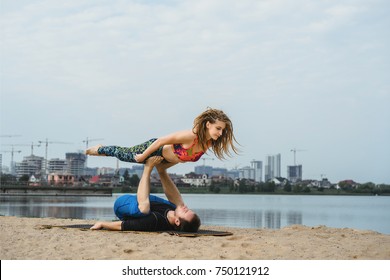 young couple practicing yoga outdoors on a city background. A girl with dreadlocks practicing hatha yoga on the shore of a lake, in the distance a big city. pair yoga man and woman, fly yoga - Powered by Shutterstock