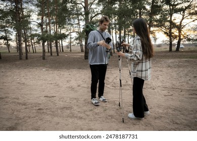 A young couple prepares their hiking gear in a forest clearing, with the woman adjusting her hiking poles and the man checking a folding chair - Powered by Shutterstock