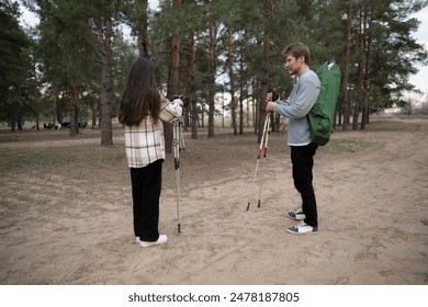 A young couple prepares their hiking gear in a forest clearing, with the woman adjusting her hiking poles and the man checking a folding chair - Powered by Shutterstock