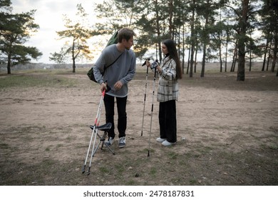 A young couple prepares their hiking gear in a forest clearing, with the woman adjusting her hiking poles and the man checking a folding chair - Powered by Shutterstock