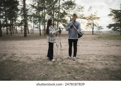 A young couple prepares their hiking gear in a forest clearing, with the woman adjusting her hiking poles and the man checking a folding chair - Powered by Shutterstock