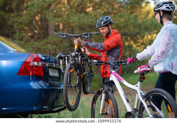 Young Couple Preparing for Riding the\
Mountain Bikes in the Forest. Unmounting the Bike from Bike Rack on\
the Car. Adventure and Family Travel\
Concept.