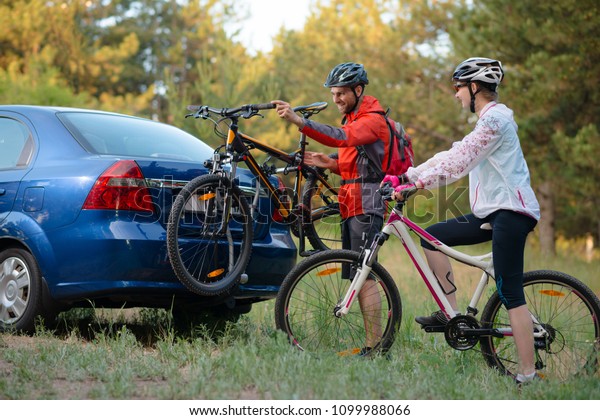Young Couple Preparing for Riding the\
Mountain Bikes in the Forest. Unmounting the Bike from Bike Rack on\
the Car. Adventure and Family Travel\
Concept.