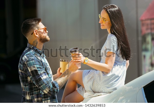 Young couple preparing for vacation trip on the\
car in sunny day. Woman and man drinking coffee and ready for going\
to sea or ocean. Concept of relationship, vacation, summer,\
holiday, weekend.