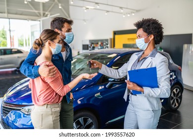 Young Couple With Protective Face Masks On Their Faces Buying New Car At Car Showroom.