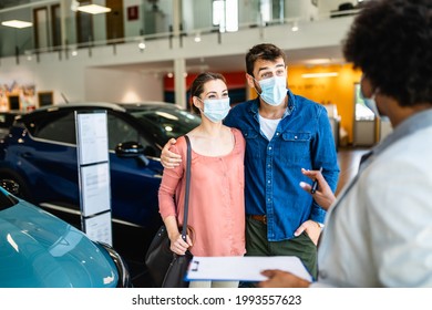 Young Couple With Protective Face Masks On Their Faces Buying New Car At Car Showroom.