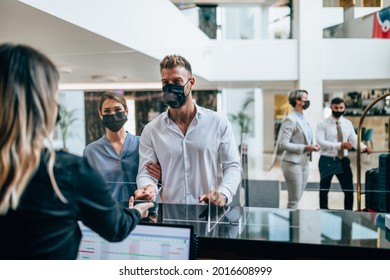 Young Couple With Protective Face Masks At The Reception Of A Hotel Checking In. Business Trip, Coronavirus, Covid-19, Safe Travel Concept. 