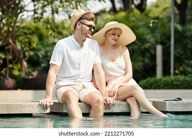 Young Couple Putting Their Feet In The Water And Enjoying Summer Vacation By The Outdoor Pool