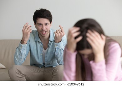 Young couple quarrelling at home, aggressive boyfriend yelling at sad lady, upset girl sitting silent holding head in hands. Mad guy accusing girlfriend of cheating. Family relationship problems 