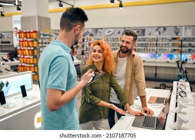 Young Couple Receives Advice From Seller In Store Of Household Appliances And Gadgets, Buying New Laptop. Male Consultant Helps In Choosing Laptop.