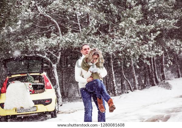 young couple in relationship hugging near yellow\
car , wearing white sweaters and blue jeans. Man roolls his woman\
on hands. The weather is snow\
