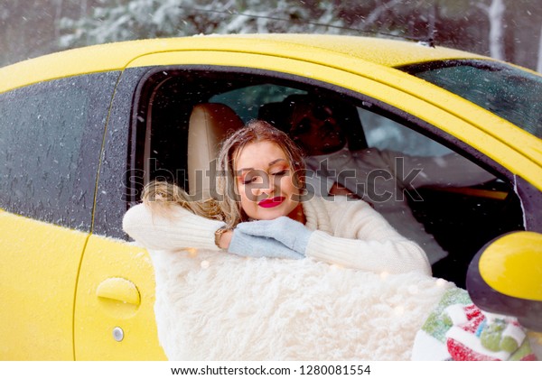 young couple in relationship hugging in yellow car ,\
wearing white sweaters and blue jeans. blond woman posing at the\
window 