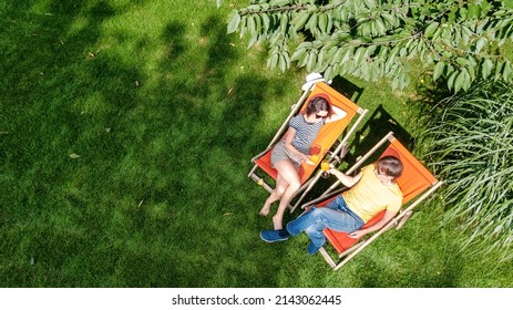 Young Couple Relax In Summer Garden In Sunbed Deckchairs On Grass, Woman And Men Have Drinks On Picnic Outdoors In Green Park On Weekend, Aerial Top View From Above