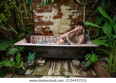 A young couple relaxes in an outdoor bubble bath among tropical plants. She playfully pushes her foot into his face.