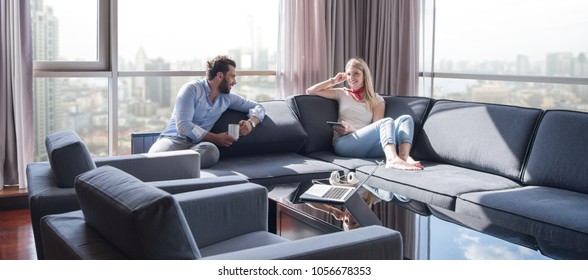 Young Couple Relaxing At  Home Using Tablet Computer Reading In The Living Room Near The Window On The Sofa Couch.